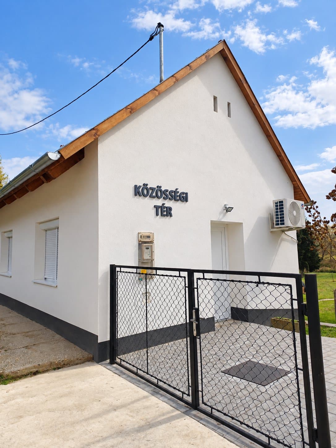 Közösségi házzá átalakított régi épület fehér homlokzattal, „Közösségi tér” felirattal és új kapuval